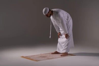 Performing the five daily prayers on time is the second pillar of Islam, drawing closer to God through the fulfillment of obligations and acts of worship. A portrait of an Emirati Gulf Arab man wearing a kandura and a white cap standing on a prayer rug in the position of bowing, with a white background.