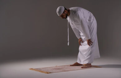 Performing the obligatory prayers on time, the second pillar of Islam, drawing closer to God through the fulfillment of duties and acts of worship, a portrait of an Emirati Arab man wearing a kandura and a white cap standing on a prayer rug in the position of bowing, white background.