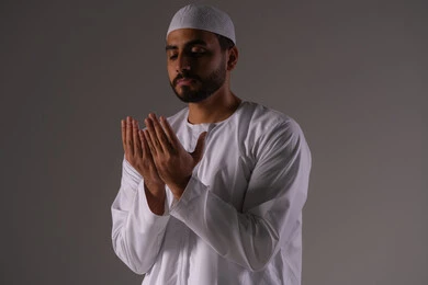 The desire to fulfill wishes, striving in worship and getting closer to the Lord of the worlds, a portrait of an Arab Gulf Emirati man wearing a kandura and a white cap, raising his hands and praying to God with gestures of supplication and humility, white background.
