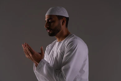 Diligence in worship and drawing closer to the Lord of the worlds, the desire to achieve wishes, a portrait of an Arab Gulf Emirati man wearing a kandura and a white cap, raising his hands and praying to God with gestures of supplication and humility, white background.