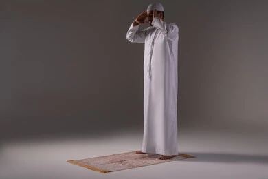 الالتزام بأداء الفروض على وقتها، ايماءات التكبير لبدء الصلاة، الركن الثاني من اركان الاسلام، بورتريه لرجل عربي خليجي إماراتي يرتدي الكندورة والقبعة البيضاء يقف على السجادة، العبادة والتقرب إلى الله، الجسم بالطول الكامل، خلفية بيضاء