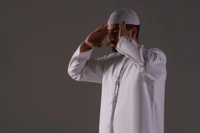 ايماءات التكبير لبدء الصلاة، الالتزام بأداء الفروض على وقتها، الركن الثاني من اركان الاسلام، بورتريه لرجل عربي خليجي إماراتي يرتدي الكندورة والقبعة البيضاء يقف على السجادة، العبادة والتقرب إلى الله، خلفية بيضاء