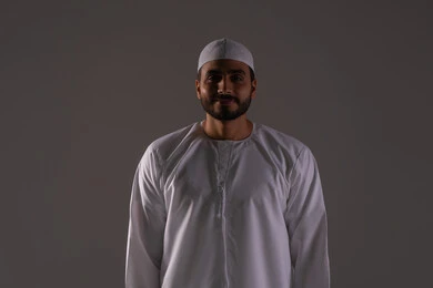 Performing Islamic religious duties, worship and drawing closer to God, establishing prayer on time, the concept of the pillars of Islam, a portrait of an Emirati Gulf Arab man wearing a kandura and a white cap standing on a prayer rug, gray background.