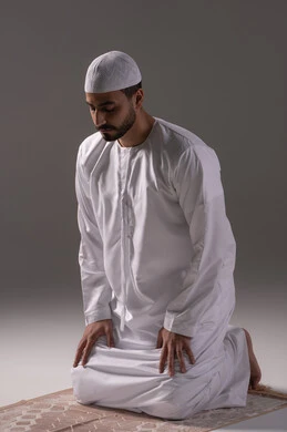 العبادة والتقرب إلى الله، الخشوع أثناء أداء فريضة الصلاة، أركان الإسلام الخمسة، بورتريه لرجل عربي خليجي إماراتي يرتدي الكندورة والقبعة البيضاء يجلس على سجادة الصلاة، خلفية رمادية