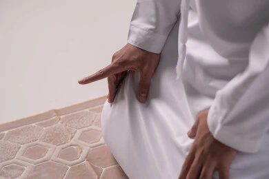 The importance of adhering to prayer on time, a close-up portrait of the hand of an Emirati Gulf Arab man wearing a kandura sitting on a prayer rug raising his index finger upwards for the testimony, worship and drawing closer to God, gray background.
