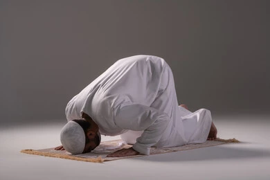 الصلاة و التضرع الى الله، مفهوم العبادة والتقرب إلى الله تعالى, بورتريه لرجل عربي خليجي إماراتي يرتدي الكندورة والقبعة البيضاء يجلس على سجادة الصلاة يقوم بالسجود ، التقرب الى الله بأداء العبادات والطاعات, خلفية رمادية