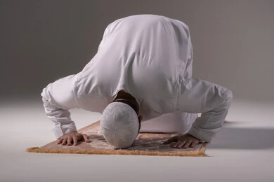 مفهوم العبادة والتقرب إلى الله تعالى, الصلاة و التضرع الى الله, بورتريه لرجل عربي خليجي إماراتي يرتدي الكندورة والقبعة البيضاء يجلس على سجادة الصلاة يقوم بالسجود ، التقرب الى الله بأداء العبادات والطاعات, خلفية رمادية
