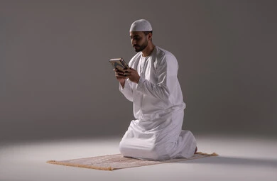 الإخلاص في العبادة والتقرب إلى الله، تلاوة وقراءة القرآن الكريم وتدبر معانيه، بورتريه لرجل عربي خليجي إماراتي يرتدي الكندورة والقبعة البيضاء يجلس على سجادة الصلاة يحمل بيده المصحف الشريف، الإكثار من ذكر الله تعالى، التقرب الى الله في شهر رمضان، الشعور بالخشوع والطمأنينة، خلفية بيضاء