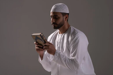 Regularly performing acts of worship, reciting and reading the Holy Quran and reflecting on its meanings, a portrait of an Emirati Gulf Arab man wearing a kandura and a white cap holding the Holy Quran in his hand, increasing the remembrance of Allah Almighty, drawing closer to Allah during the month of Ramadan, feeling humility and tranquility, white background.
