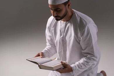 Reading the Quran with contemplation and reflection, feeling humility and tranquility, a portrait of an Emirati Gulf Arab man wearing a kandura and a white cap reading the Holy Quran, increasing the remembrance of Allah through seeking forgiveness and prayer, striving in worship, drawing closer to Allah during the month of Ramadan, white background.