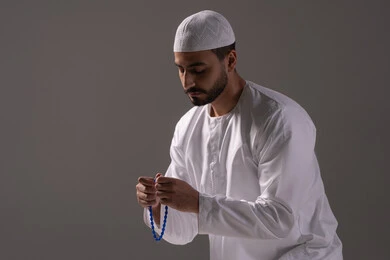 الإكثار من الأذكار اليومية وذكر الله، العبادة والطاعة والتقرب الى الله، بورترية لشاب عربي خليجي اماراتي يرتدي الثوب الأبيض والطاقية يمسك بيده سبحة من حبات من الخرز الزرقاء ، الاجتهاد في العبادات، خلفية رمادية