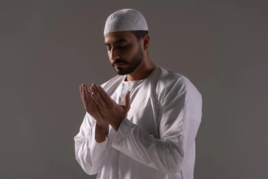The feeling of humility and tranquility, the remembrance of Allah Almighty with abundant supplication and seeking forgiveness, a portrait of a young Arab Gulf Emirati man wearing a white thobe and a cap, raising his hands and praying to Allah with humility and supplication, drawing closer to the Lord of the Worlds in the blessed month of Ramadan, striving in worship and performing religious duties, with a gray background.