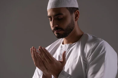 The mention of Allah, along with abundant supplication and seeking forgiveness, is depicted in a close-up portrait of a young Arab Gulf Emirati man wearing a white thobe and a cap, raising his hands and praying to Allah with humility and earnestness, drawing closer to the Lord of the Worlds during the blessed month of Ramadan, striving in worship and performing religious duties, with a gray background.
