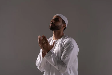 الالتزام بالعبادات والفرائض اليومية، الاستغفار والإكثار من الدعاء وذكر الله تعالى، بورترية لشاب عربي خليجي اماراتي يرتدي الثوب الأبيض والطاقية يرفع يديه ويدعو الله بخشوع وتضرع، التقرب الى رب العالمين في شهر رمضان المبارك، خلفية رمادية
