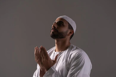 The commitment to obligatory acts and worship, seeking forgiveness and increasing supplication and remembrance of Allah Almighty, a close-up portrait of a young Emirati Arab man wearing a white thobe and a cap, raising his hands and praying to Allah with humility and supplication, drawing closer to the Lord of the Worlds in the blessed month of Ramadan, with a gray background.