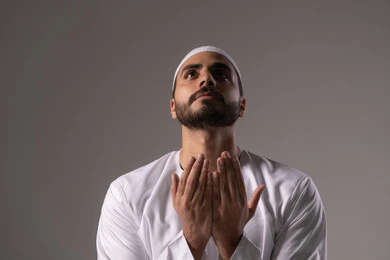 Diligence in acts of worship and religious obligations, seeking forgiveness and increasing supplication and remembrance of Allah Almighty, a close-up portrait of a young Emirati Arab man wearing a white thobe and a cap, raising his hands and praying to Allah with humility and supplication, drawing closer to the Lord of the Worlds in the blessed month of Ramadan, with a gray background.