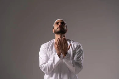 الالتزام بالعبادة وفريضة الدين، الاستغفار والإكثار من الدعاء وذكر الله تعالى، بورترية لشاب عربي خليجي اماراتي يرتدي الثوب الأبيض والطاقية يرفع يديه ويدعو الله بخشوع وتضرع، التقرب الى رب العالمين في شهر رمضان المبارك، خلفية رمادية