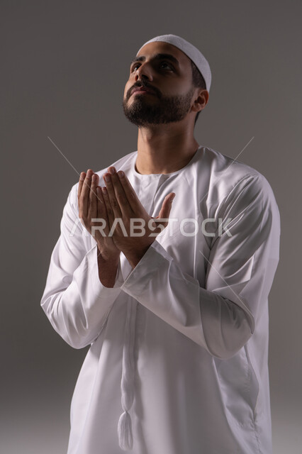 الحرص على أداء الصلوات والواجبات الدينية، الاستغفار والإكثار من الدعاء وذكر الله تعالى، بورترية لشاب عربي خليجي اماراتي يرتدي الثوب الأبيض والطاقية يرفع يديه ويدعو الله بخشوع وتضرع، التقرب الى رب العالمين في شهر رمضان المبارك، خلفية رمادية