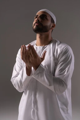 The commitment to performing prayers and religious duties, seeking forgiveness, and increasing supplication and remembrance of Allah Almighty, a portrait of a young Emirati Arab man wearing a white thobe and a cap, raising his hands and praying to Allah with humility and supplication, drawing closer to the Lord of the Worlds in the blessed month of Ramadan, with a gray background.