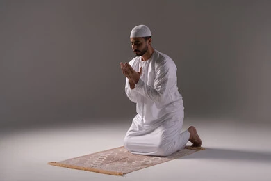 المواظبة على العبادات والفرائض، الاستغفار والإكثار من الدعاء وذكر الله تعالى، بورترية لشاب عربي خليجي اماراتي يرتدي الثوب الأبيض والطاقية يجلس على سجادة الصلاة يرفع يديه ويدعو الله بخشوع وتضرع، التقرب الى رب العالمين في شهر رمضان المبارك، خلفية رمادية