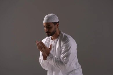الاجتهاد في العبادة واداء الفرائض الدينية، التقرب الى رب العالمين في شهر رمضان المبارك، الاستغفار والإكثار من الدعاء وذكر الله تعالى، بورترية لشاب عربي خليجي اماراتي يرتدي الثوب الأبيض والطاقية يجلس على سجادة الصلاة يرفع يديه ويدعو الله بخشوع وتضرع، خلفية رمادية