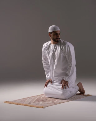 The concept of the pillars of Islam, performing prayer on time, fulfilling Islamic religious obligations, an Emirati Arab man wearing a kandura and a white cap sitting on a prayer rug performing the tasleem at the end of his prayer, the feeling of humility and tranquility, worship and drawing closer to God, white background.