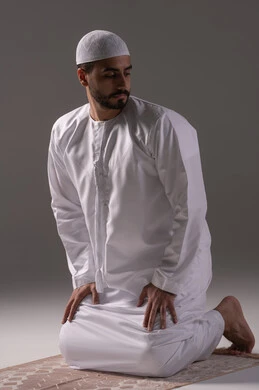 إقامة الصلاة على وقتها، أداء الفرائض الدينية الإسلامية, مفهوم أركان الإسلام، رجل عربي خليجي إماراتي يرتدي الكندورة والقبعة البيضاء يجلس على سجادة الصلاه يقوم بالتسليم في ختام صلاته، الشعور بالخشوع والطمأنينة، العبادة والتقرب إلى الله، خلفية بيضاء