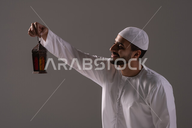 الإستعداد لقدوم شهر رمضان المبارك ، أجواء السكينة والروحانية، الأعياد والمناسبات الدينية، بورترية لرجل عربي خليجي إماراتي يرتدي الكندورة البيضاء والطاقية يحمل بيدية فانوس معدني ينظر إليه، العبادة والتقرب إلى الله، خلفية رمادية