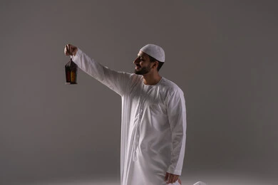 The atmosphere of tranquility and spirituality, holidays and religious occasions, a portrait of an Emirati Gulf Arab man wearing a white kandura and a cap, holding a metal lantern in his hands and looking at it, preparing for the arrival of the blessed month of Ramadan, worship and drawing closer to God, with a gray background.