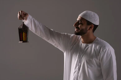 الأعياد والمناسبات الدينية، أجواء السكينة والروحانية، بورترية لرجل عربي خليجي إماراتي يرتدي الكندورة البيضاء والطاقية يحمل بيدية فانوس معدني ينظر إليه، الإستعداد لقدوم شهر رمضان المبارك ، العبادة والتقرب إلى الله، خلفية رمادية