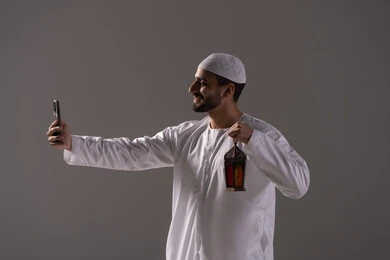 قضاء أوقات روحانية في شهر رمضان المبارك، التقاط الصور لفانوس رمضان، العبادة والتقرب إلى الله، بورترية من الجانب لرجل عربي خليجي إماراتي يرتدي الكندورة البيضاء والطاقية يمسك بيده الهاتف المحمول، خلفية رمادية قضاء أوقات روحانية في شهر رمضان المبارك، التقاط الصور لفانوس رمضان، العبادة والتقرب إلى الله، بورترية من الجانب لرجل عربي خليجي إماراتي يرتدي الكندورة البيضاء والطاقية يمسك بيده الهاتف المحمول، خلفية رمادية