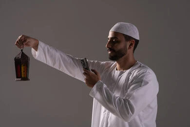 التقاط الصور لفانوس رمضان، العبادة والتقرب إلى الله، الاستمتاع بالأجواء الروحانية في الشهر المبارك، بورترية من الجانب لرجل عربي خليجي إماراتي يرتدي الكندورة البيضاء والطاقية يمسك بيده الهاتف المحمول، خلفية رمادية
