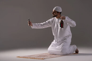 الاستمتاع بالأجواء الروحانية في شهر رمضان المبارك، بورترية لرجل عربي خليجي إماراتي يرتدي الكندورة البيضاء والطاقية  يجلس على سجادة الصلاة يمسك بيده الهاتف المحمول يقوم بالتقاط صورة لفانوس معدني، العبادة والتقرب إلى الله، خلفية رمادية