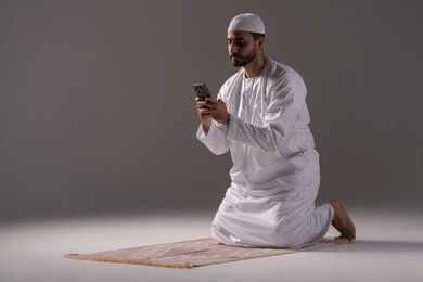 Browsing smart applications, communicating online, a portrait of an Arab Gulf Emirati man wearing a white kandura and a head cap sitting on a prayer rug using a mobile phone with gestures of engagement and focus, using modern technologies, gray background. Browsing smart applications, communicating online, a portrait of an Arab Gulf Emirati man wearing a white kandura and a head cap sitting on a prayer rug using a mobile phone with gestures of engagement and focus, using modern technologies, gray background.