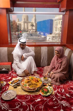 قضاء الأوقات الممتعة في الخارج، ايماءات السعادة و السرور، مفهوم الترابط الأسري، مطاعم و كافيهات في الإمارات، مائدة مليئة بتشكيلة متنوعة من المأكولات, رجل عربي خليجي اماراتي يرتدي الكندورة و الغتره يجلس فى مجلس ارضي شعبي برفقة زوجته