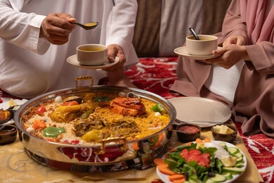 مائدة مليئة بتشكيلة متنوعة من المأكولات، قضاء الأوقات الممتعة في الخارج, صورة مقربة لرجل عربي خليجي اماراتي يرتدي الكندورة يقوم بتناول صحن من الشوربة برفقة زوجته , مطاعم و كافيهات في الإمارات، الإجتماع العائلي في المناسبات الدينيه السعيده