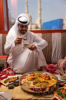 قضاء الأوقات الممتعة في الخارج، سفره مليئة بتشكيلة متنوعة و مختلفة من المأكولات, رجل عربي خليجي اماراتي يرتدي الكندورة و الغتره يجلس فى مجلس ارضي شعبي يقوم بتناول صحن من الشوربة، تعابير السعادة و السرور, مطاعم و كافيهات في الإمارات