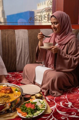 سفره مليئة بتشكيلة متنوعة و مختلفة من المأكولات, امراه عربيه خليجيه اماراتية ترتدي العباءة و الحجاب تجلس فى مجلس ارضي شعبي تقوم بتناول صحن من الشوربة، تعابير السعادة و السرور, مطاعم و كافيهات في الإمارات, قضاء الأوقات الممتعة في الخارج