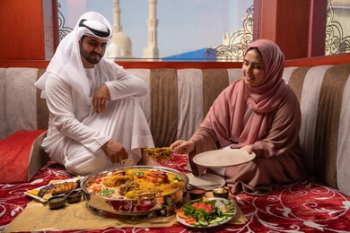 اجتماع الأسرة حول مائدة الطعام، أجواء دينية رمضانية، إمراة عربية خليجية اماراتية ترتدي العباءة والحجاب تقوم بوضع الطبق الرئيسي، سفرة افطار مليئة باصناف من المأكولات الشهية في شهر رمضان، أداء فريضة الصيام، إيماءات السعادة و السرور