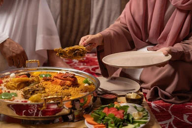 Ramadan religious atmosphere, a close-up image of the hand of an Emirati Gulf Arab woman placing the main dish, an iftar table filled with a variety of delicious foods during the month of Ramadan, family gathering around the dining table, observing the fasting obligation, gestures of happiness and joy. Ramadan religious atmosphere, a close-up image of the hand of an Emirati Gulf Arab woman placing the main dish, an iftar table filled with a variety of delicious foods during the month of Ramadan, family gathering around the dining table, observing the fasting obligation, gestures of happiness and joy.