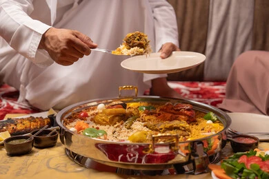 سفرة مليئة باصناف من المأكولات اللذيذة،  تناول وجبة الافطار، صورة مقربة ليد  رجل عربي خليجي اماراتي يرتدي الكندورة على مائدة الافطار بشهر رمضان، موعد الإفطار عند اذان المغرب