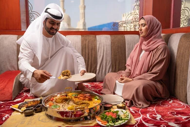 Performing the duty of fasting, Ramadan religious atmosphere, an Emirati Gulf Arab man wearing a kandura and ghutrah is placing the main dish alongside his wife, an iftar table filled with a variety of delicious foods during the month of Ramadan, family gathering around the dining table, gestures of happiness and joy.