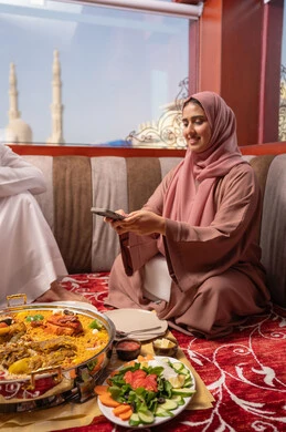 Preserving joyful memories, gestures of happiness and joy, an Emirati Gulf Arab family dressed in traditional attire sitting in a popular floor seating area around the dining table, the concept of family bonding, taking commemorative photos of food with a mobile phone.