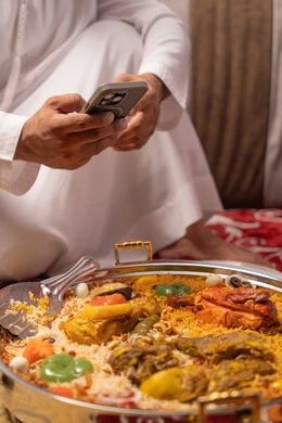 توثيق اللحظات السعيدة ، تصوير مقرب ليد رجل عربي خليجي اماراتي يرتدي الكندورة و الغتره يجلس فى مجلس ارضي شعبي حول مائدة الطعام ، التقاط  صور تذكارية  للمائدة عبر الهاتف المحمول