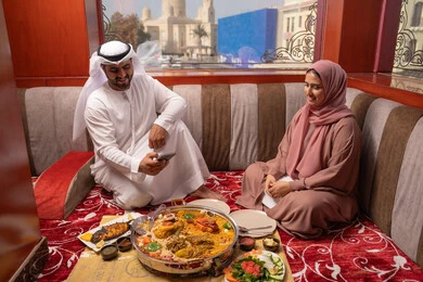 Capturing special moments, gestures of happiness and joy, an Emirati Gulf Arab family dressed in traditional attire sitting in a popular floor seating area around the dining table, the concept of family bonding, taking commemorative photos of the food with a mobile phone.