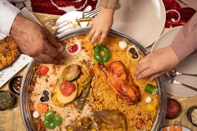 A rich breakfast with a variety of dishes and appetizers, delicious healthy recipes, a metal tray filled with rice and spices, a close-up image of the hands of an Emirati Gulf Arab family enjoying a plate of rice garnished with stuffed chicken.