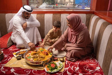 A Ramadan table filled with delicious food and appetizers, restaurants and cafes in the United Arab Emirates, an Emirati Gulf Arab family dressed in traditional attire sitting in a popular floor seating area around the dining table.