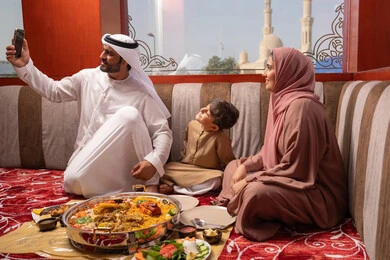 Gestures of happiness and joy, an Emirati Gulf Arab family dressed in traditional attire sitting in a popular ground council around a dining table, the concept of family bonding, taking selfies with a mobile phone, documenting happy memories.