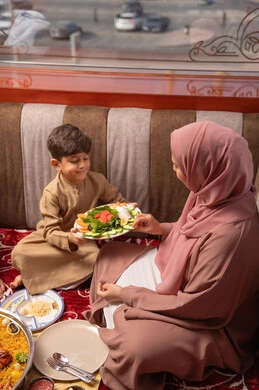 A mother's care for her children, a breakfast spread filled with a variety of delicious foods and appetizers, strengthening the bond of motherhood between the mother and her son, an Arab Gulf Emirati woman wearing an abaya and hijab sitting in a traditional floor seating area, giving her son a plate of healthy salad, gestures of happiness and joy.