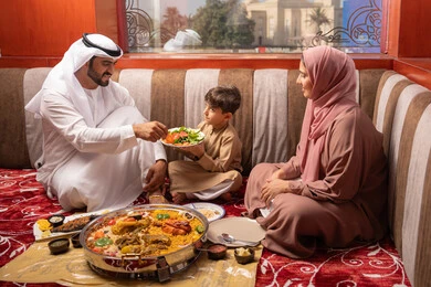 مائدة إفطار مليئة بالمأكولات والمقبلات الشهية، أجواء شهر رمضان المبارك ، قضاء وقت ممتع فى الخارج، رجل عربي خليجي اماراتي يرتدي الكندورة و الغتره يجلس فى مجلس ارضي شعبي يقوم بتناول الافطار مع ابنه  و زوجته ، ايماءات السعادة و السرور